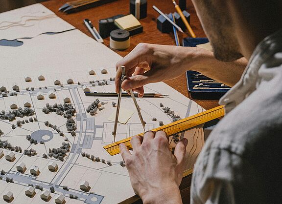 Ein Mann im Hemd ist von hinten zu sehen und zeichnet mit Lineal und Zirkel etwas auf einen Stadtplan.