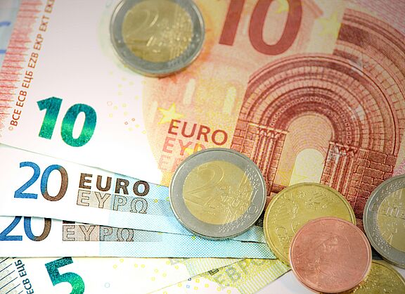Es sind mehrere Geldscheine zu sehen. Ein Zehn-Euro-Schein liegt auf mehreren Zwanzig-Euro- und einem Fünf-Euro-Schein. Außerdem liegen einige Münzen auf den Scheinen verstreut.
