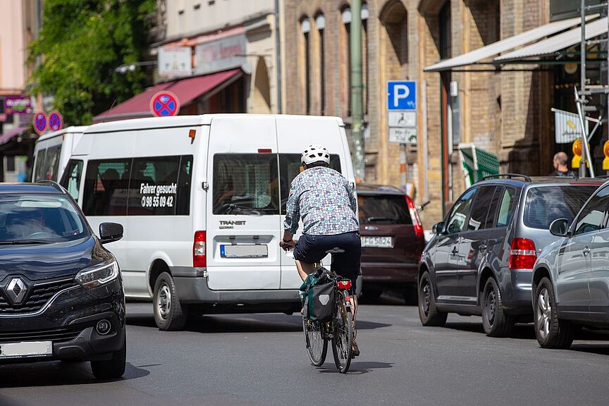 Radfahrer auf Geschäftsstraße neben parkenden Autos.