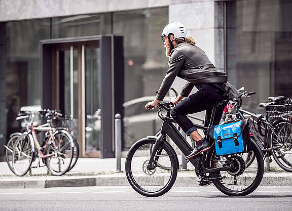 Berufspendler in der Stadt Ein Mann mit Bart und weißem Helm fährt auf einem modernen Elektrofahrrad durch eine städtische Straße. Er trägt Alltagskleidung (Jacke und Jeans) und hat eine leuchtend blaue Packtasche am Gepäckträger befestigt. Im Hintergrund sind Gebäude mit großen Glasfronten und parkende Fahrräder unscharf zu erkennen.